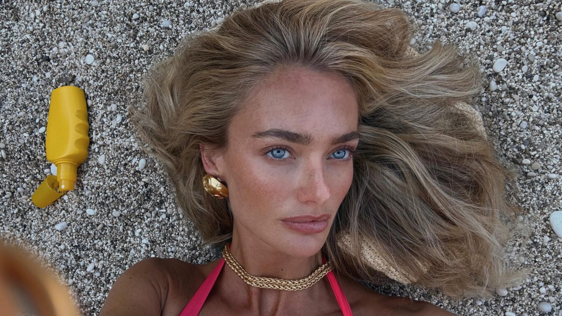 Chica rubia con bikini rojo y joyas doradas en la playa con un protector solar