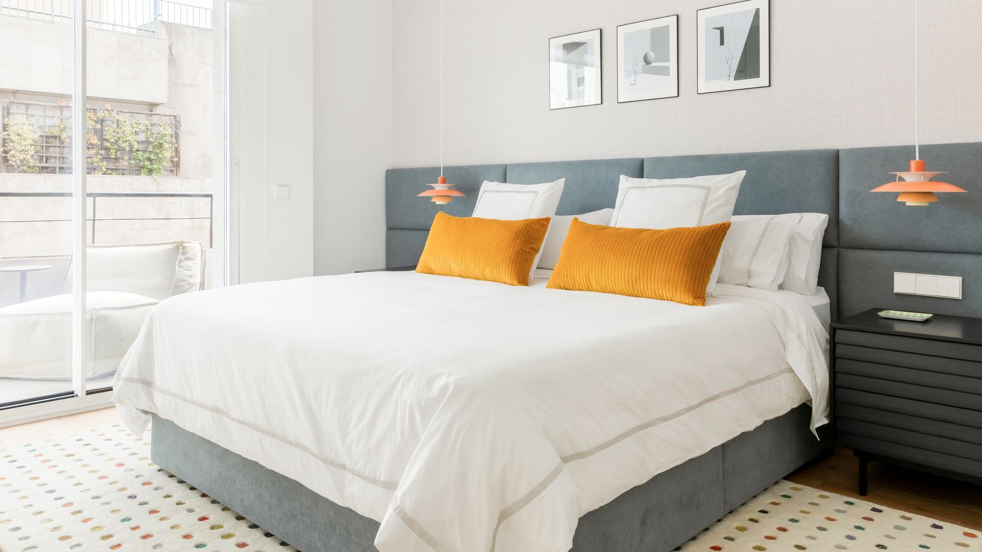 Dormitorio con cabecero tapizado que ocupa todo el ancho de la pared, ventanal con salida a la terraza, cuadros sobre la cama, ropa de cama blanca y cojines amarillos, lámparas de techo a ambos lados de la cama
