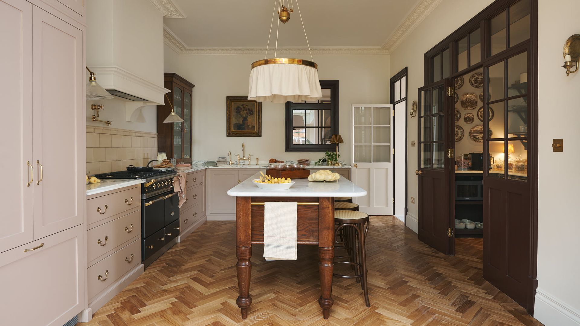 Cocina con mesa de madera y sobre de mármol en el centro, taburetes, suelo de madera en espiga, muebles de cocina rosas, lámpara de techo, despensa con platos en la pared