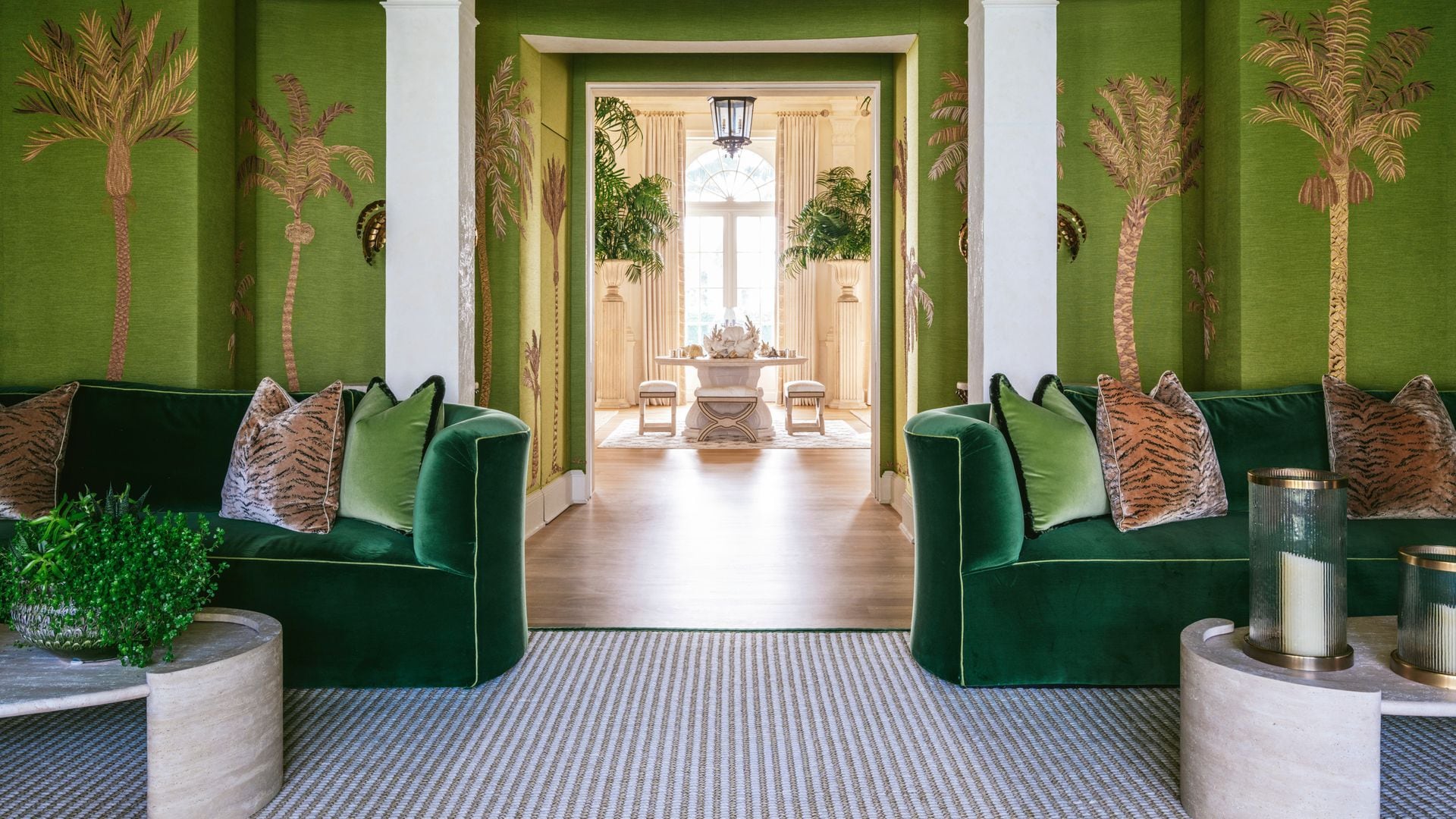 Sala verde con sofás verdes en los laterales, cojines, dos mesas blancas con plantas y pared con decoración tropical de palmeras en amarillo