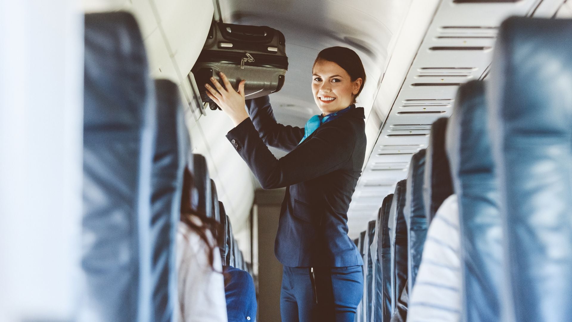 Azafata en cabina de avión colocando una maleta en la parte superior.