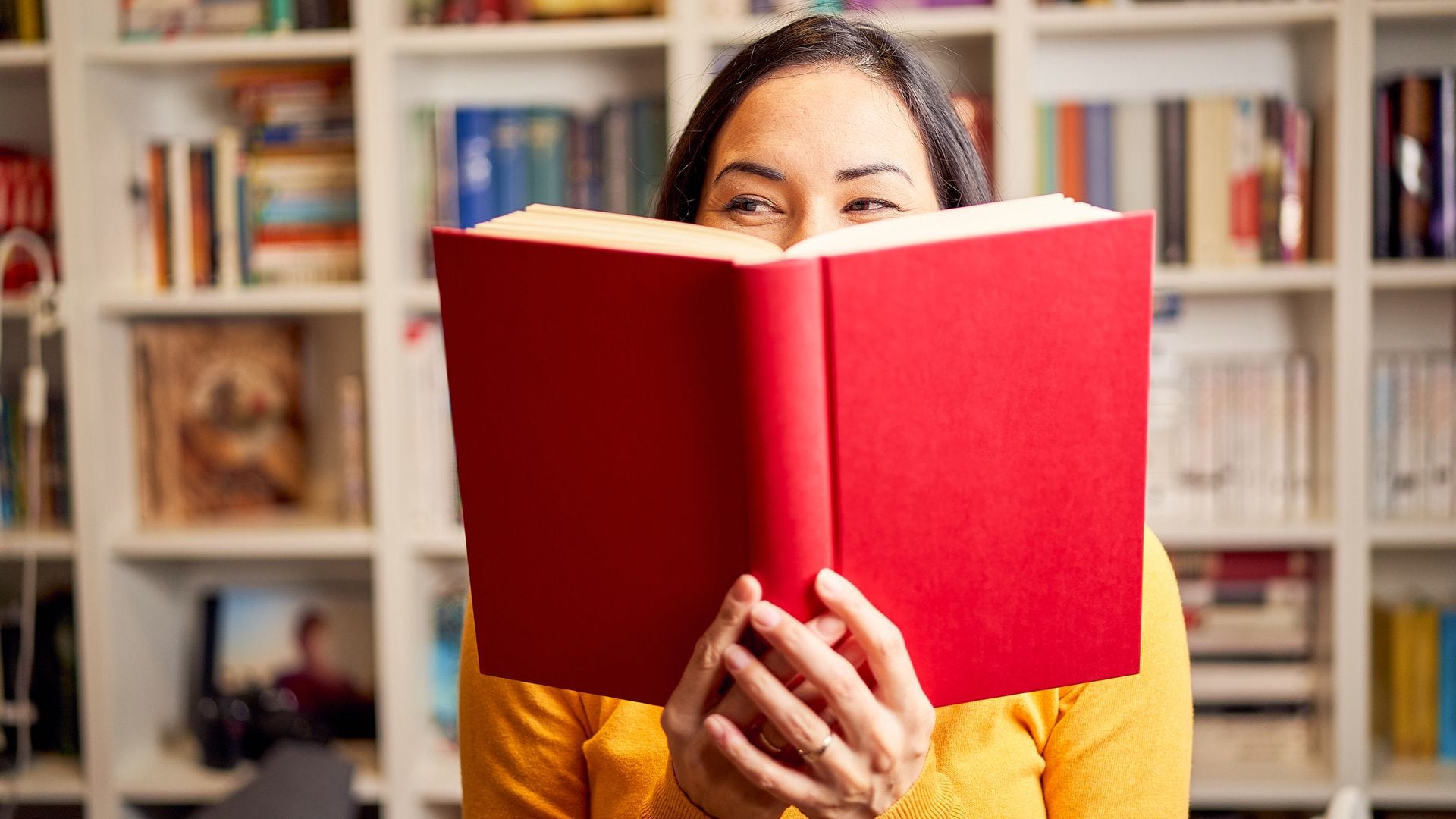 Mujer sonriendo mirando hacia un lado mientras sostiene un libro de cubierta roja entre las manos. Detrás tiene una estantería repleta de libros.