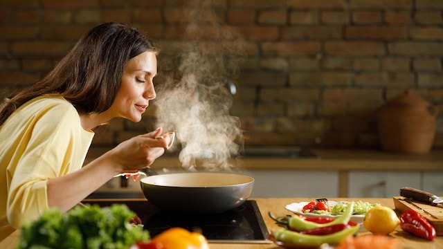 Mujer cocinando en la cocina, oliendo una receta que tiene en el fuego en una sartén tipo 'wok'. En la encimera, hay una tabla de cortar y muchas verduras.
