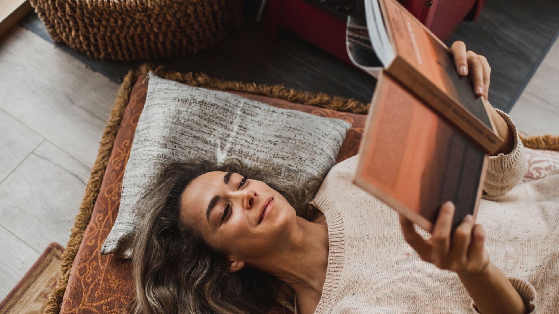 Mujer leyendo un libro tumbada en el suelo, sobre unos cojines y alfombra en tonos marrones y verdes. Lleva jersey y el pelo suelo. Está tumbada cerca de una chimenea.