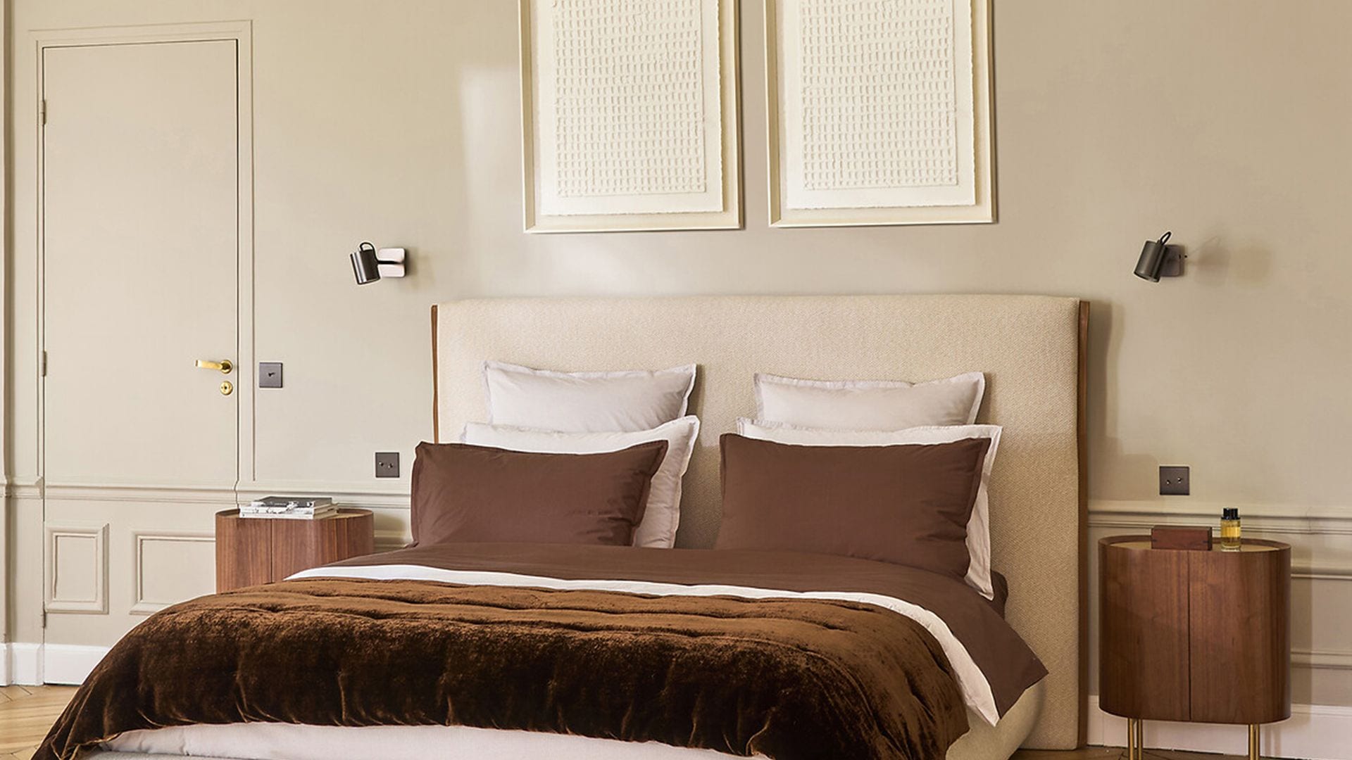 Dormitorio con paredes, puerta y molduras en color beige, cabecero tapizado a juego, cuadros en la pared del cabecero, alfombra, apliques de pared, mesitas de noche con patas doradas, manta suave de color marrón oscuro y cojines marrones