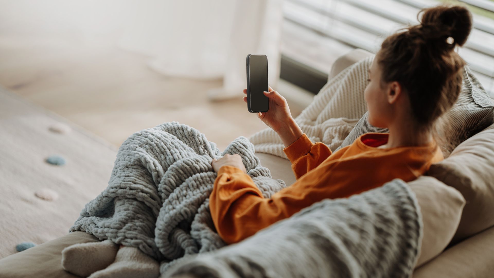 Mujer joven en casa tapada con manta en el sofá, con el teléfono móvil en la mano y ropa de estar por casa.