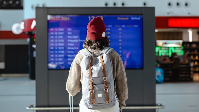 Mujer mirando una pantalla de llegadas y salidas en tren o aeropuerto con una mochila en invierno