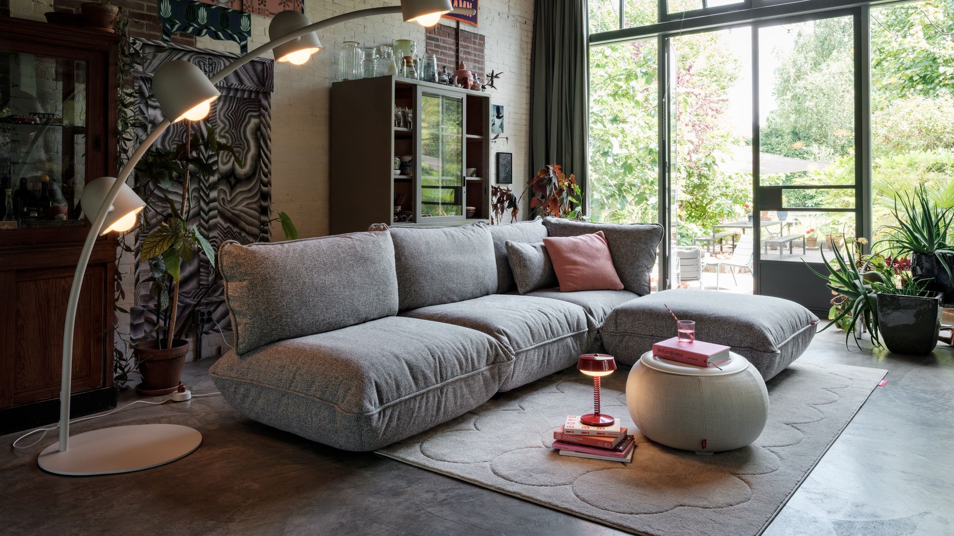 Salón con sofá modular gris, salida al jardín, lámpara de pie, estantería vitrina, alfombra, puf