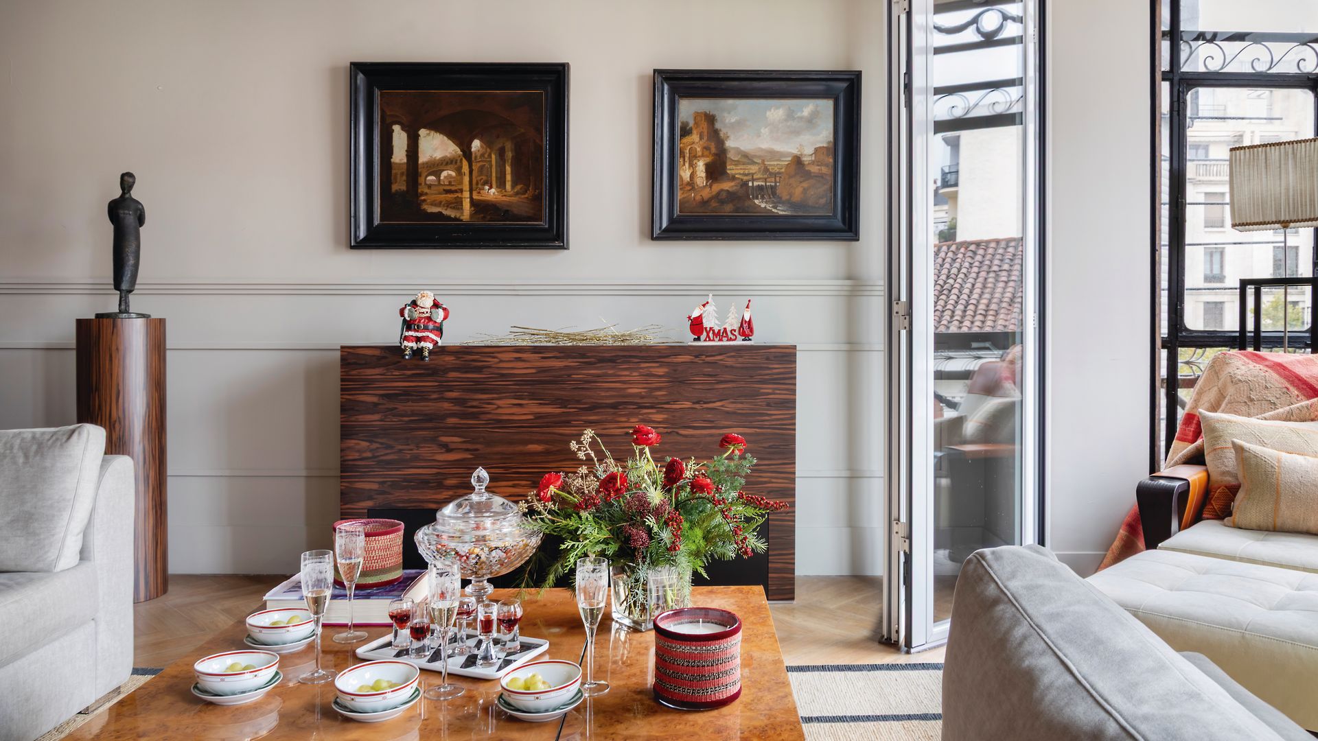 Salón con decoración navideña., mesa con centro de flores rojo, copas, velas de color rojo, adornos navideños sobre el mueble, cuadros en la pared