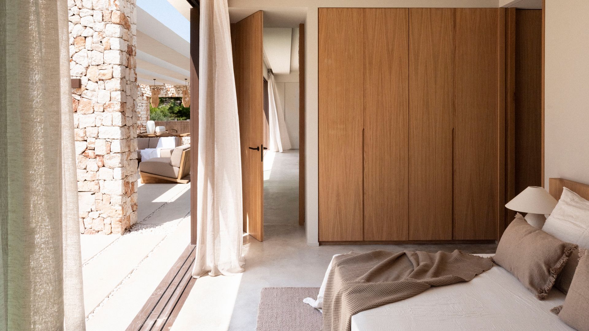 Dormitorio abierto al jardín con armario de madera, vigas en el techo y cortinas de lino beige