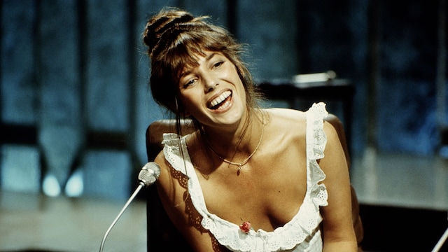 Plano medio de Jane Birkin con top blanco de volantes y jeans azules sentada