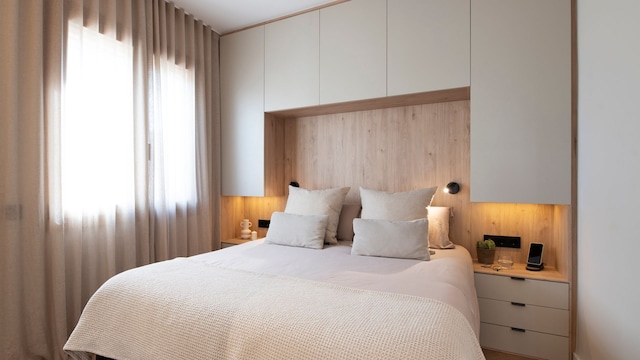 Dormitorio en tonos neutros con cortinas beiges y estructura puente sobre la cama que integra las mesitas de noche y una zona de armarios en la parte superior