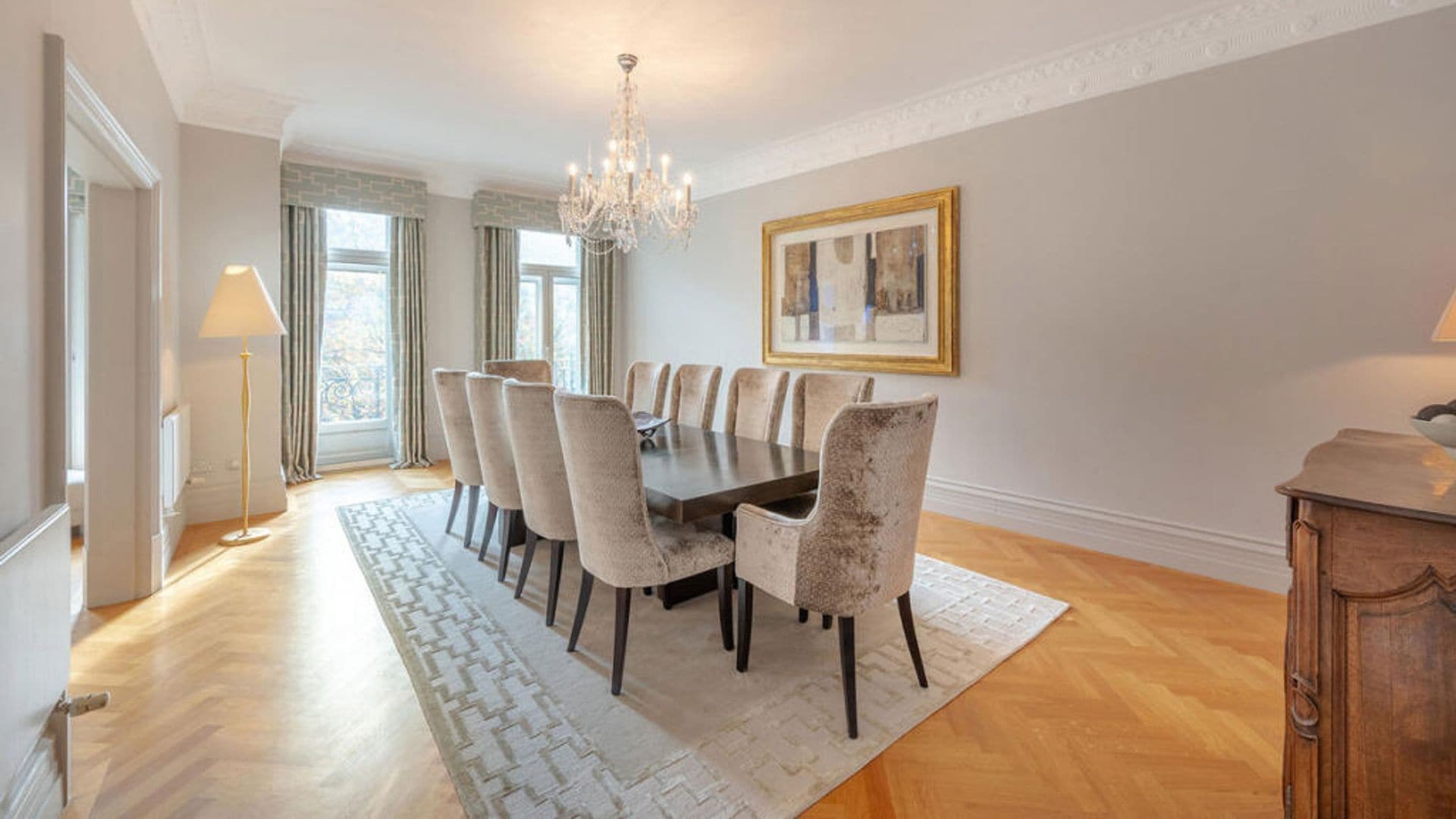 Comedor de estilo clásico con gran mesa rectangular, alfombra, lámpara de araña, cuadros y sillas tapizadas
