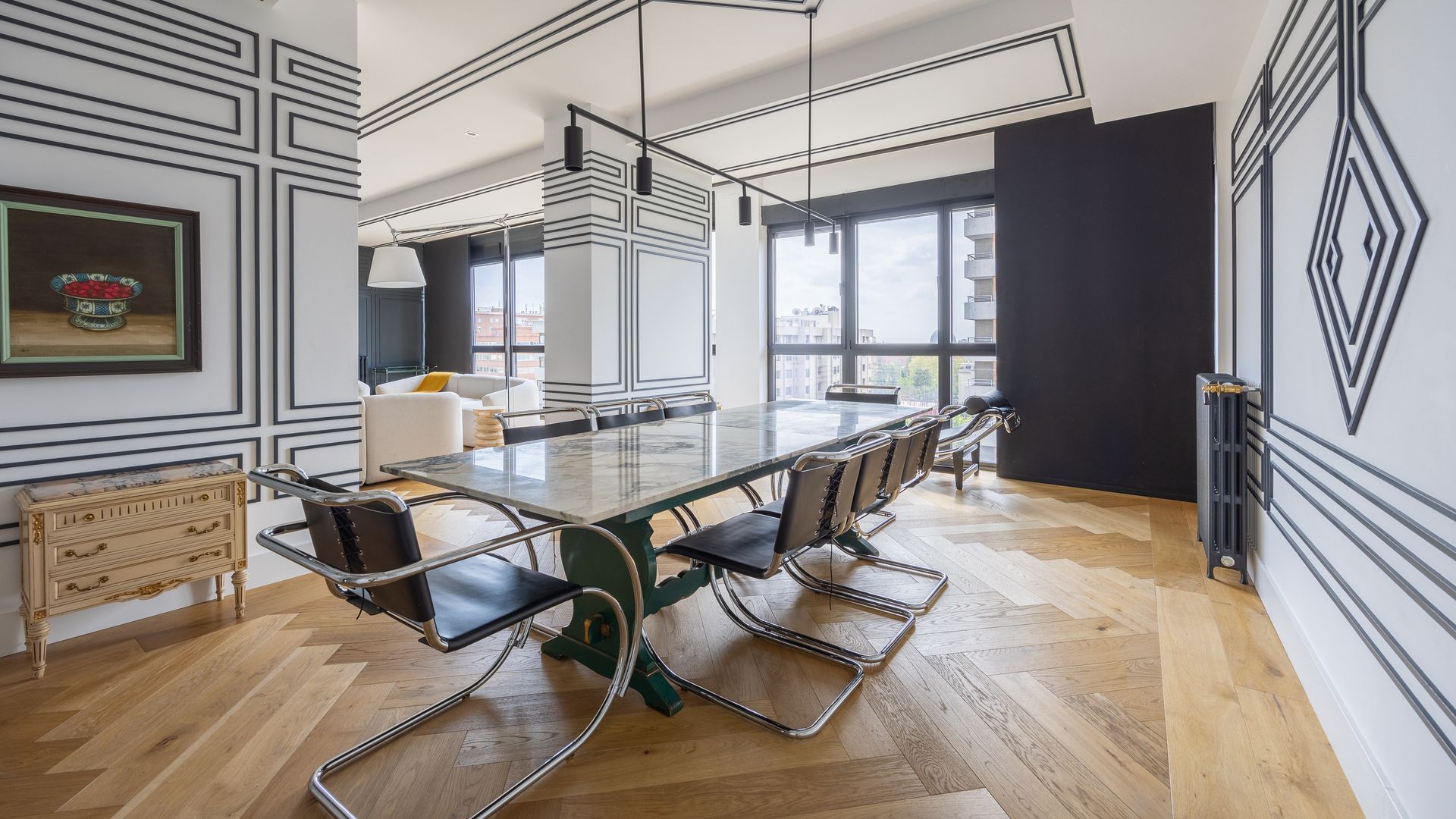 Comedor con mesa XL, sillas metálicas y cuero negro, paredes y techos blancos con molduras decorativas azules, suelo de madera, cuadro en la pared