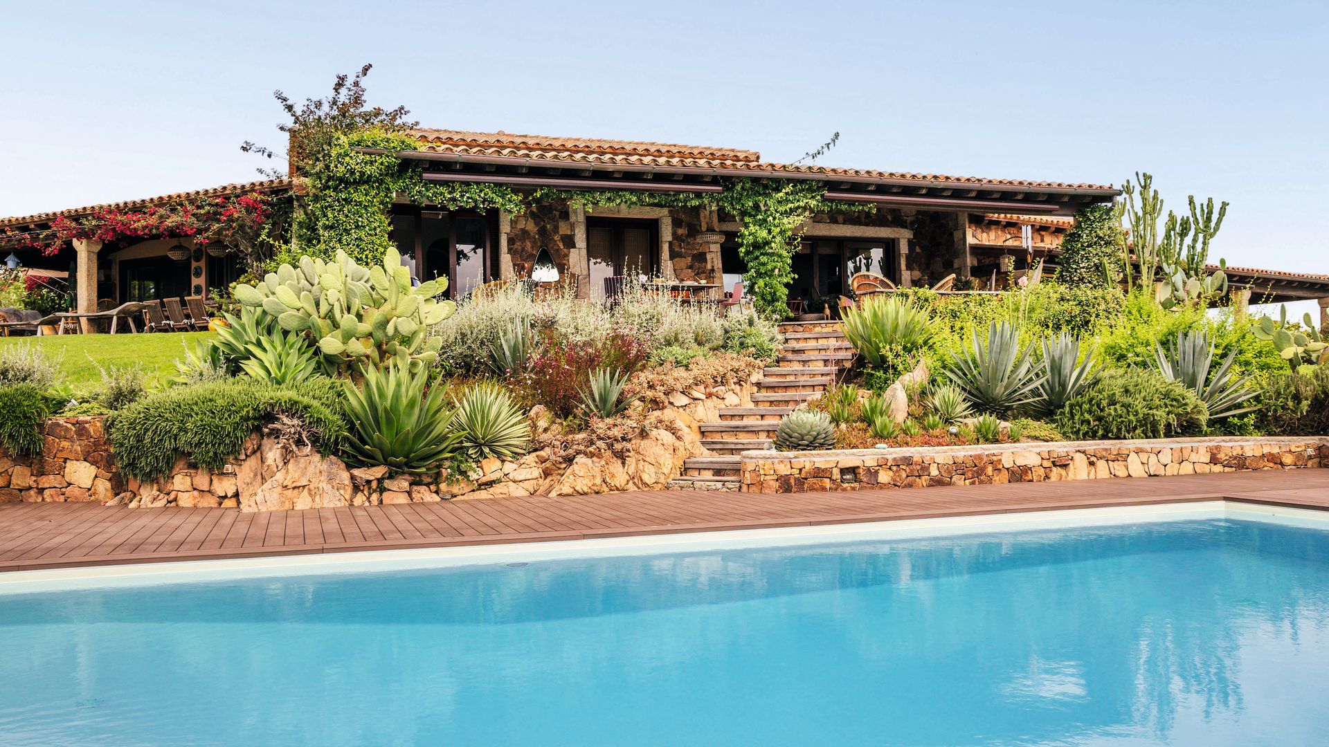Una vista de  la villa, con fachada de piedra, que se asoma al mar entre terrazas, jardines y una piscina que parece fundirse con el horizonte