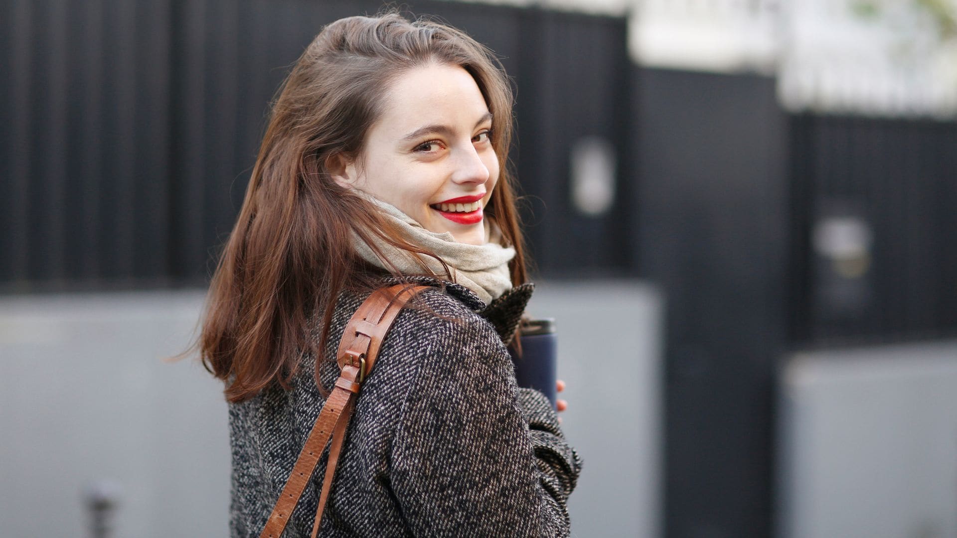 Mujer joven sonriendo hacia la cámara, mirando hacia detrás. Lleva el pelo liso, labios rojos y va con ropa de invierno, con un bolso colgado al hombro mientras camina por la calle.
