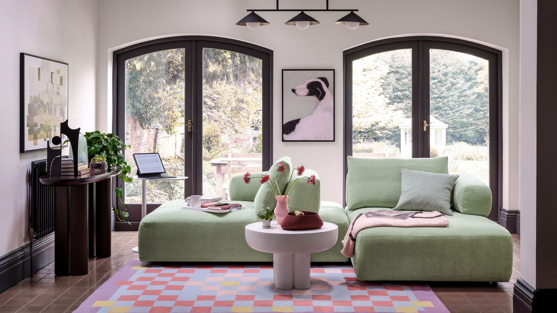 Salón con sofás modulares en color verde claro, alfombra rosa, ventanales con salida al jardín, mesa de centro rosa, lámpara de techo, consola de madera oscura