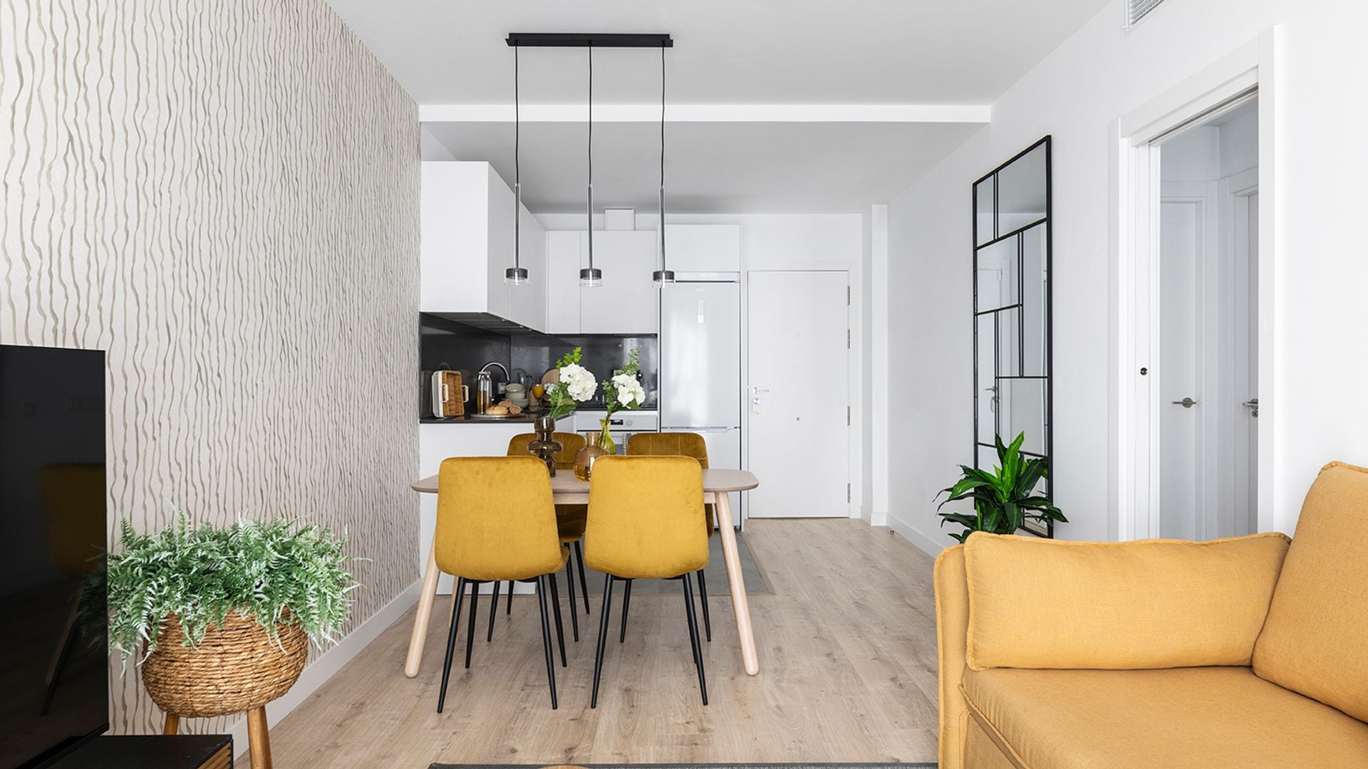 Salón, comedor cocina en uno, con entrada directa desde la calle, suelo de madera, sofá mostaza a juego con las sillas, pared de espejo y lámpara de techo