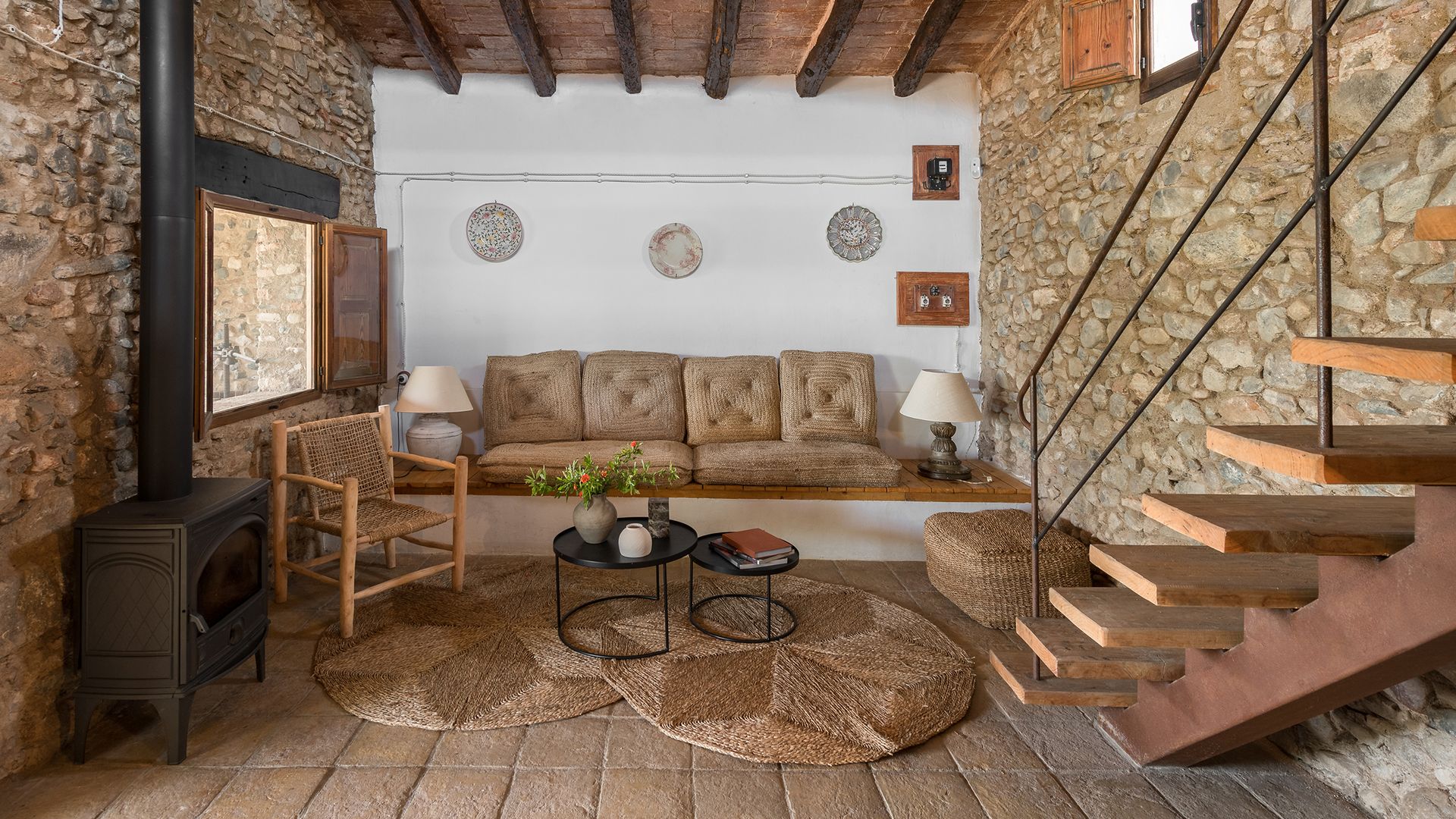 Salón de estilo rústico, de casa de campo con sofá de obra, techo con vigas, alfombra de fibras vegetales redonda, platos en la pared, chimenea, paredes de piedra
