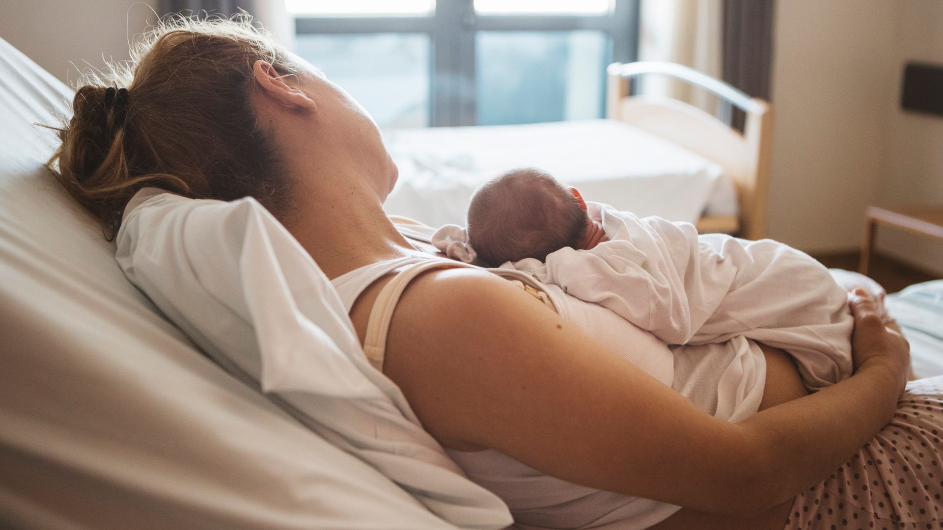 Una madre con su bebé recién nacido tumbados en la cama de un hospital mirando hacia la ventana.