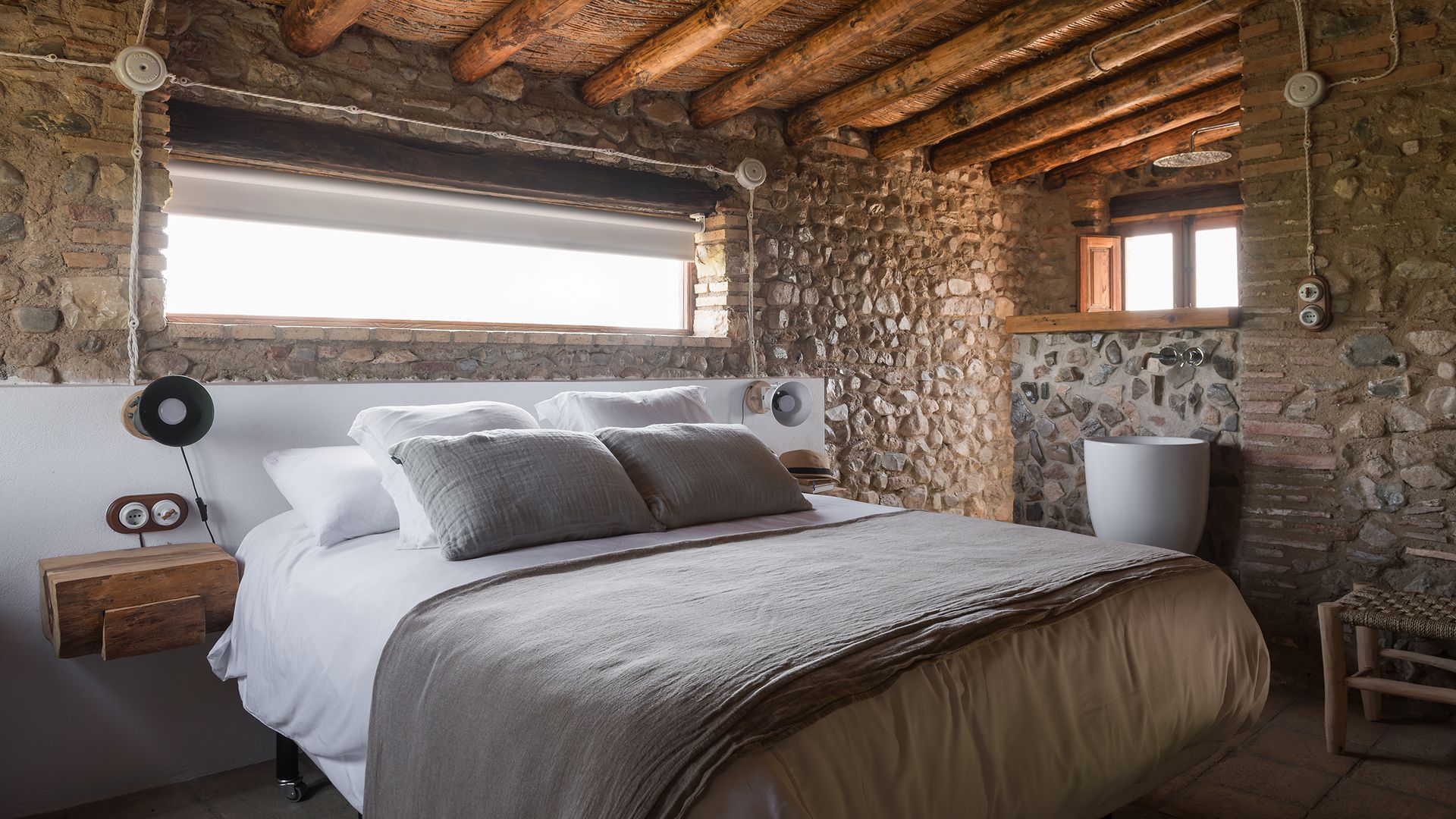 Dormitorio con vigas de madera en el techo, paredes de piedra, baño integrado, ventana horizontal