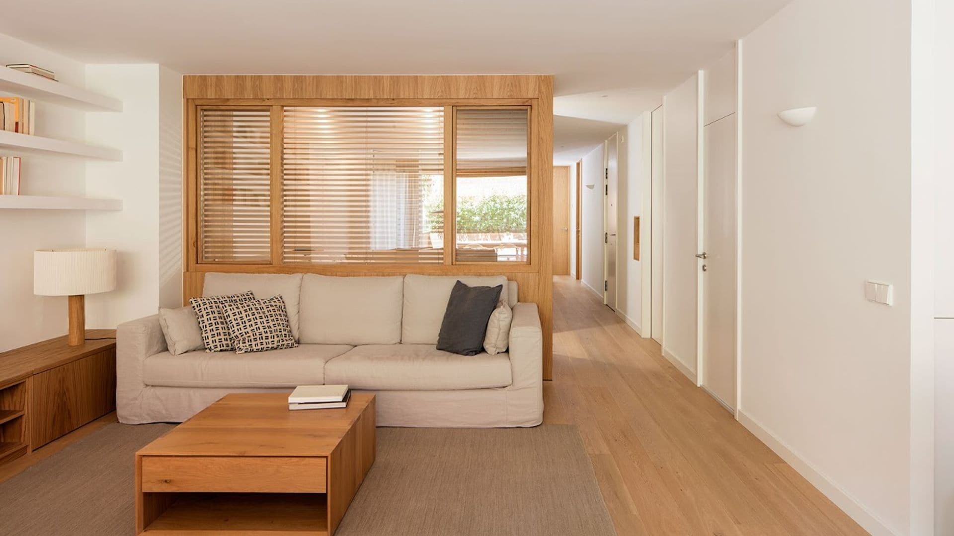 Salón con tabiqué de madera abierto separando el estar del comedor, mesa de centro acierta en los materiales, sofá beige, baldas en la pared, estantería baja con puertas 