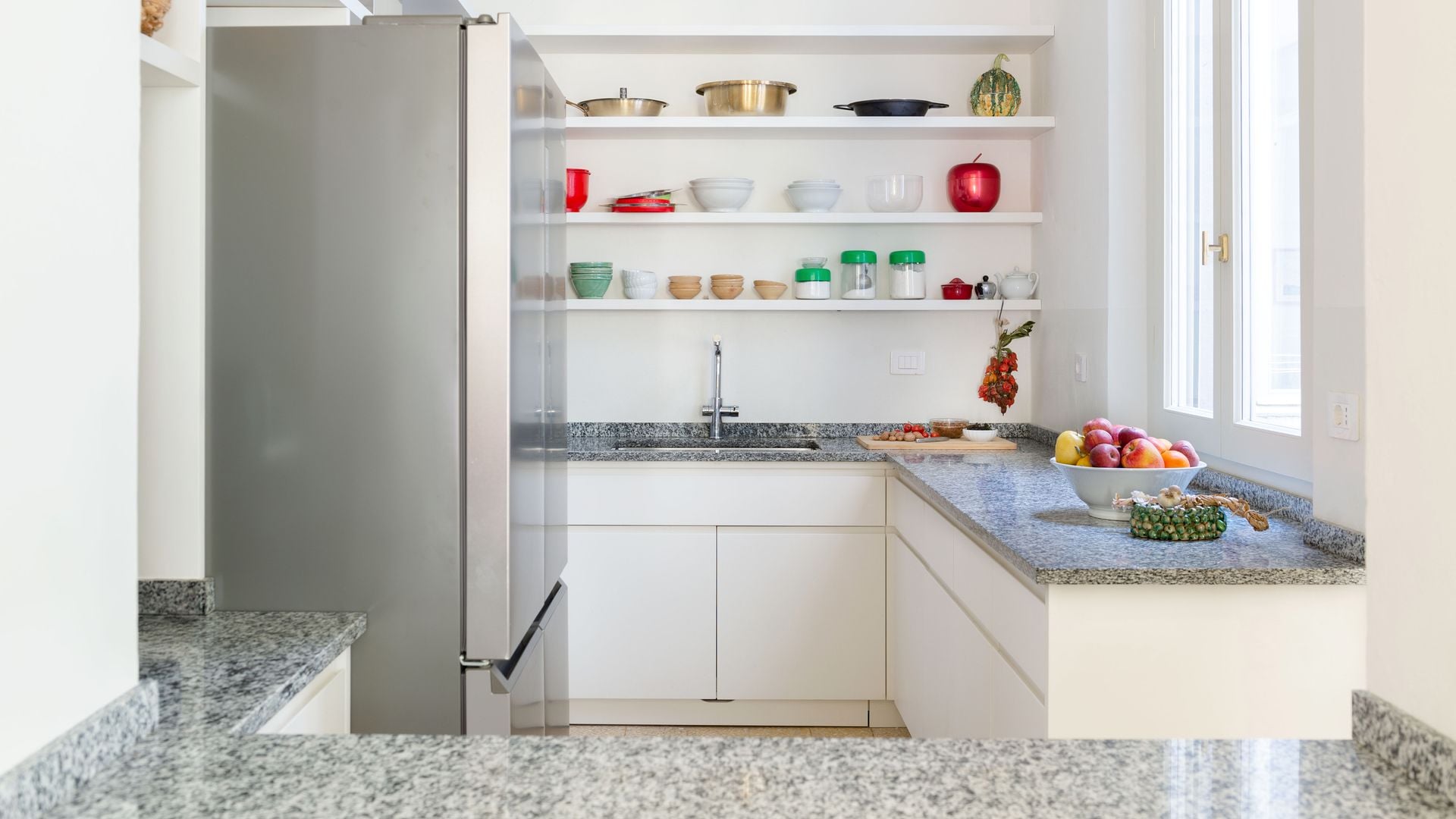 Cocina blanca con encimera de granito, sin armarios superiores, con baldas en la parte superior, frigorífico XL de acero inoxidable, ventanas de madera 