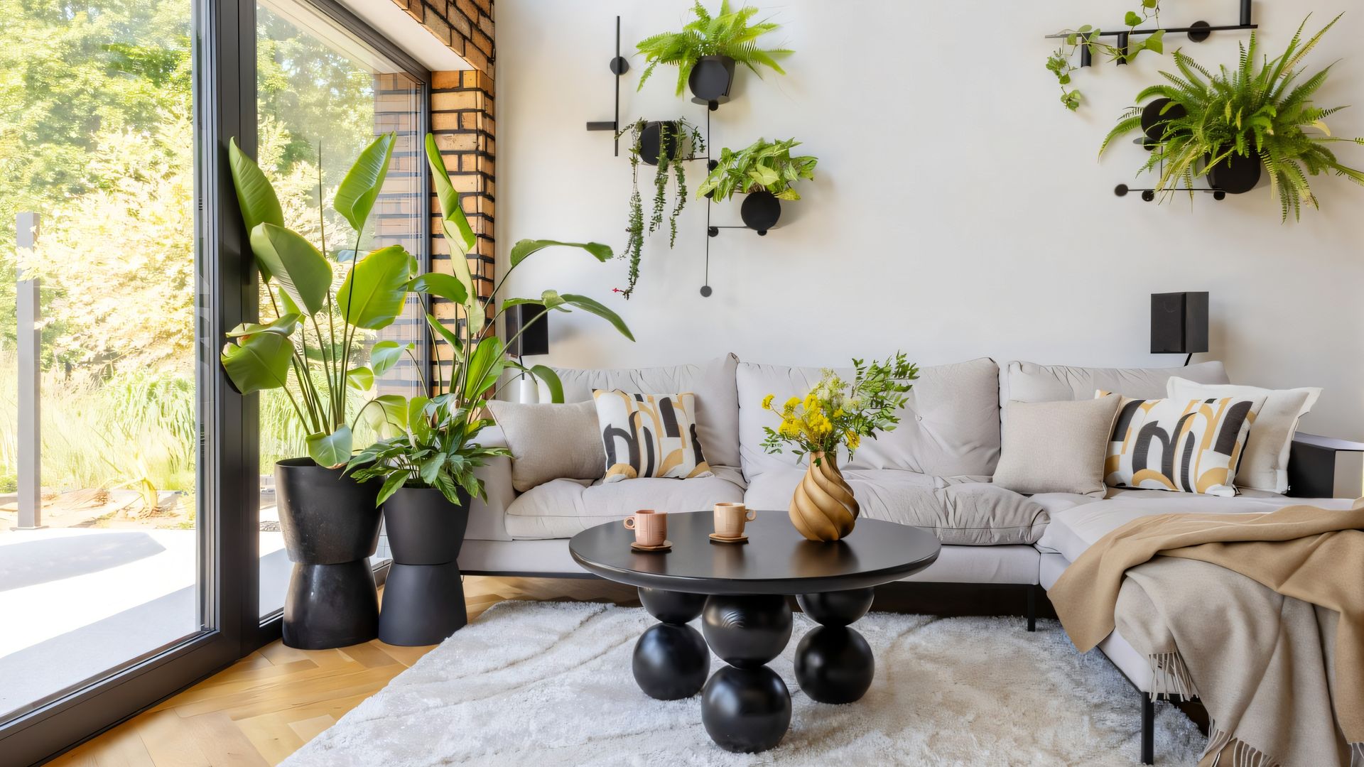 Salón con salida al jardín, sofá beige, alfombra, mesa de centro negra, plantas colgantes y plantas XL en el suelo en macetas negras