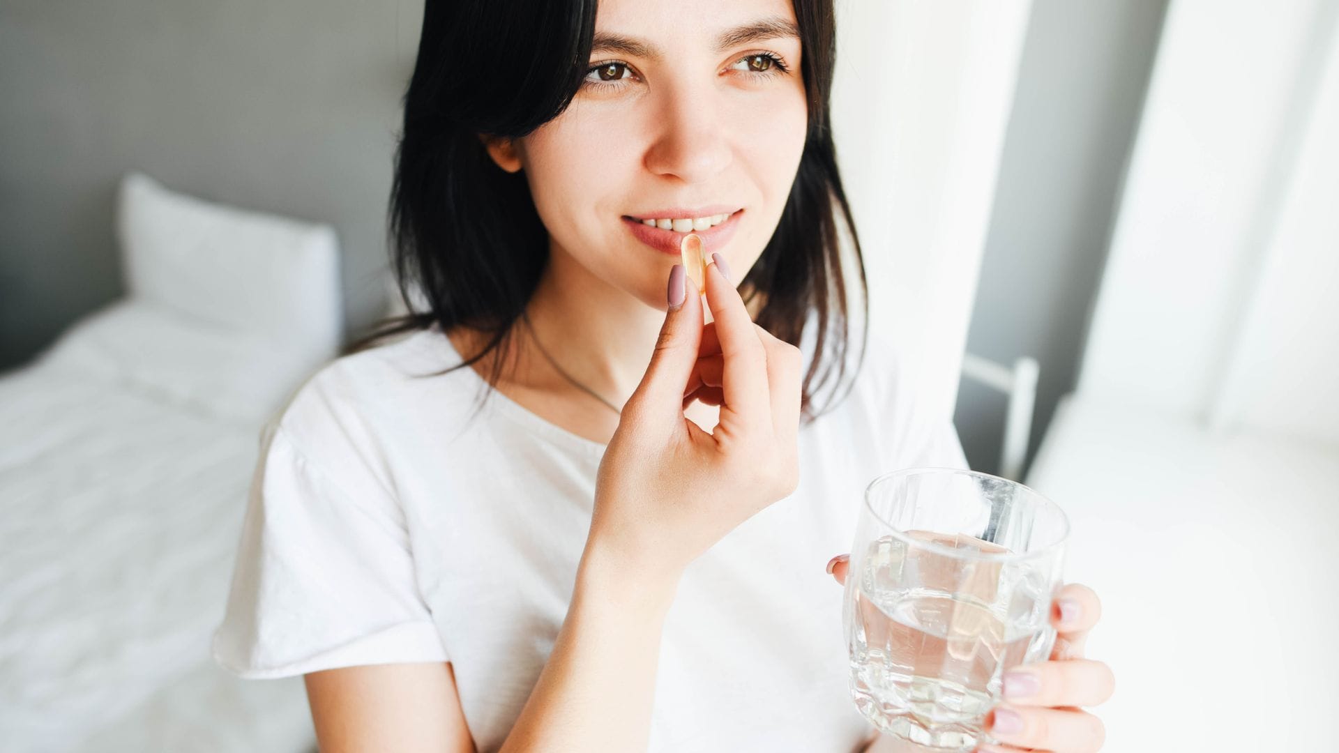 Mujer de mediana edad tomando un suplemento alimenticio con un vaso de agua en la mano. Está en un dormitorio, cama blanca y ella también va vestida de blanco.