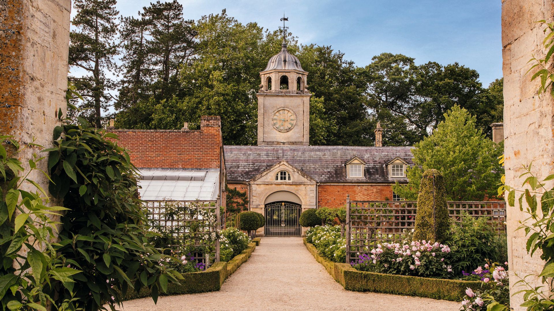 Vista de un jardín con camino de grava, flores, y una casa con torre y reloj.