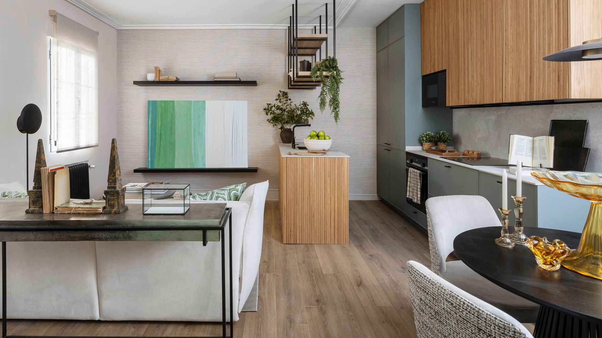 Espacio único con salón, comedor y cocina, muebles de cocina grises y de madera, consola de hierro y cristal, estor blanco, cuadro en la pared