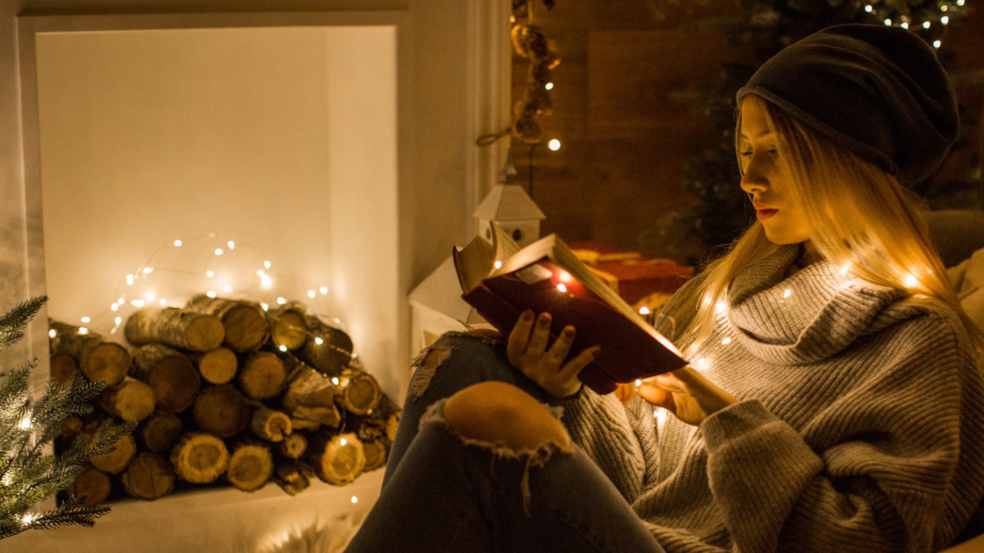 Chica joven leyendo un libro en Navidad, cerca de una chimenea, con luces navideñas por el salón.