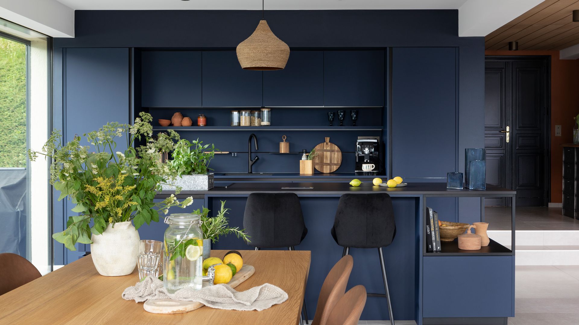 Cocina con isla abierta al comedor en color azul fuerte, con taburetes negros, lámpara de techo de fibras vegetales