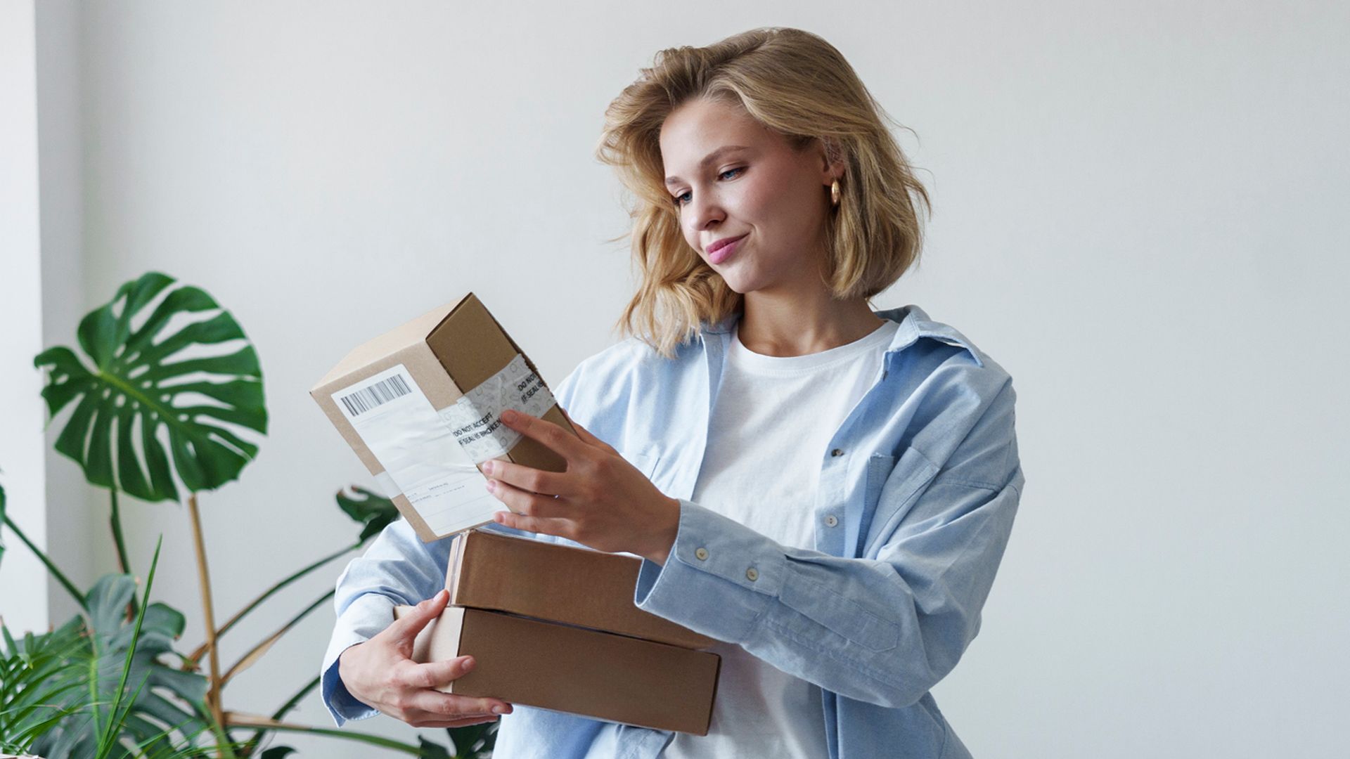 Mujer rubia con camisa azul, vaqueros y camiseta blanca revisando cajas y pedidos en casa