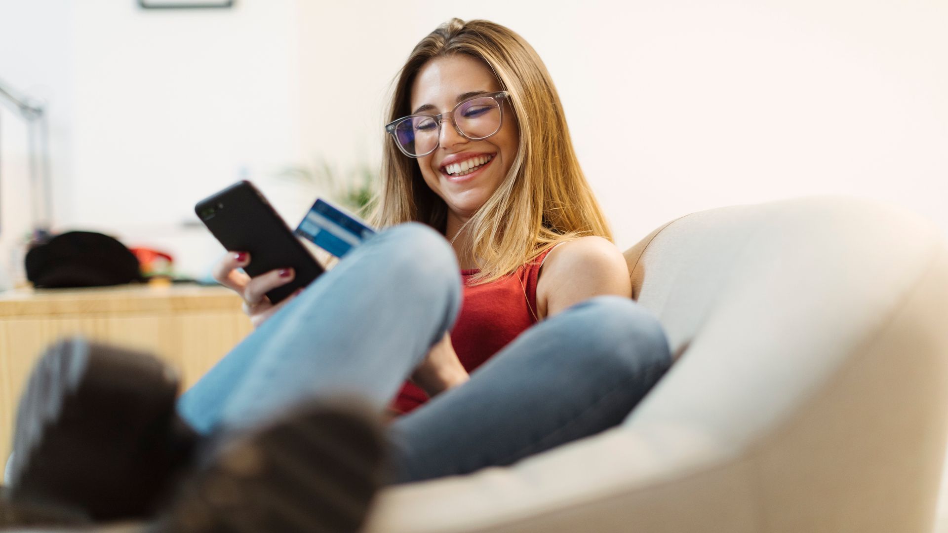 Mujer joven sonriente con un teléfono móvil en la mano y una tarjeta de crédito haciendo compras, con ropa de verano, sentada en el sofá.