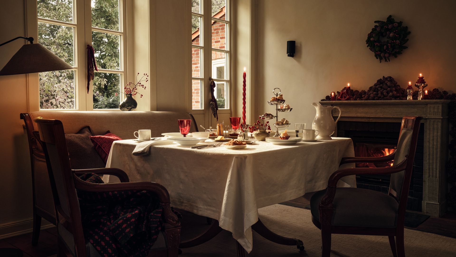 Comedor con chimenea, banco, mesa de Navidad, mantel blanco, candelabro rojo, vajilla de porcelana blanca, chimenea con decoración, corona navideña sobre la chimenea, grandes ventanales