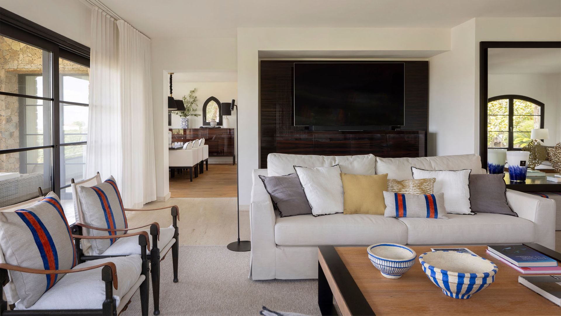 Salón con sofás blanco, televisión en mueble, lámpara de pie negra, cojines, sillas de madera con cojines, alfombra, vista del comedor
