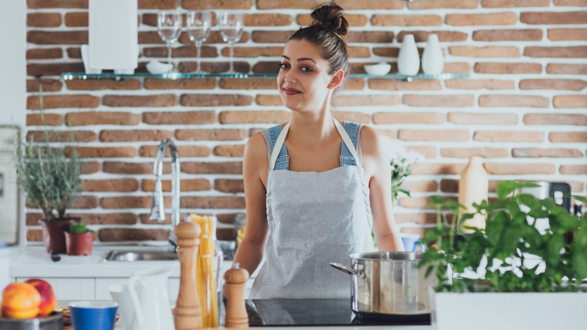 Mujer cocinando detrás de una isla de cocina con una cacerola en la vitrocerámica. Tiene spaguettis, especias y otros ingredientes. El fondo es de ladrillo visto, con el fregadero y estaterías.