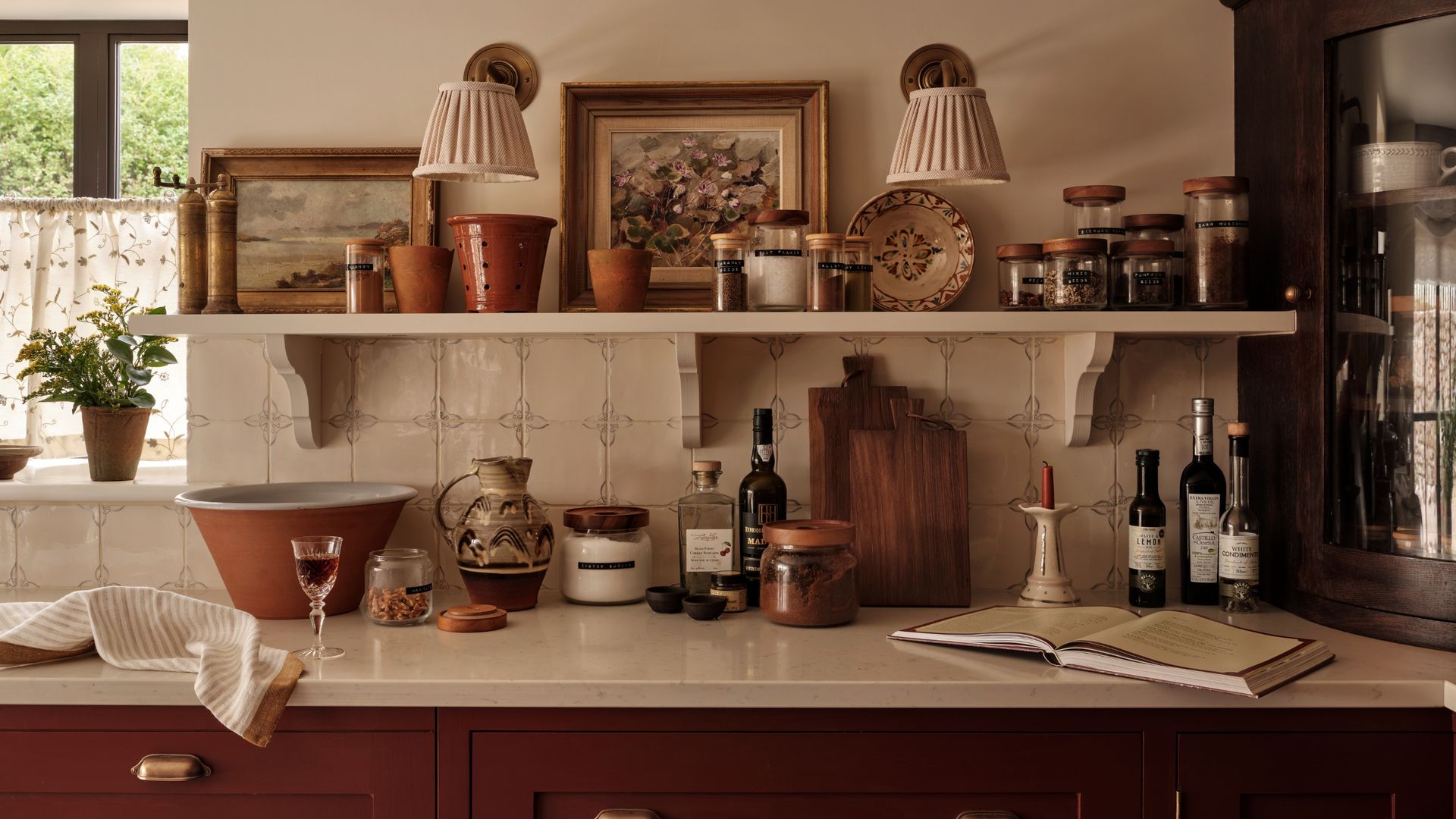 Cocina cottage con azulejos, balda de madera blanca, muebles de madera oscura, apliques con la pantalla de tela, botes y tarros de especies, libro de recetas