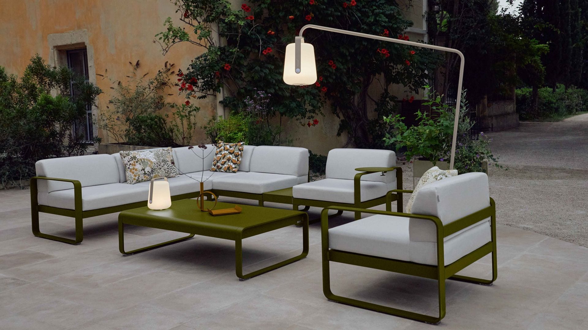 Salón exterior con conjunto de mesa, sofá y butacas verdes con almohadones grises, lámpara de pie y de sobremesa portátiles
