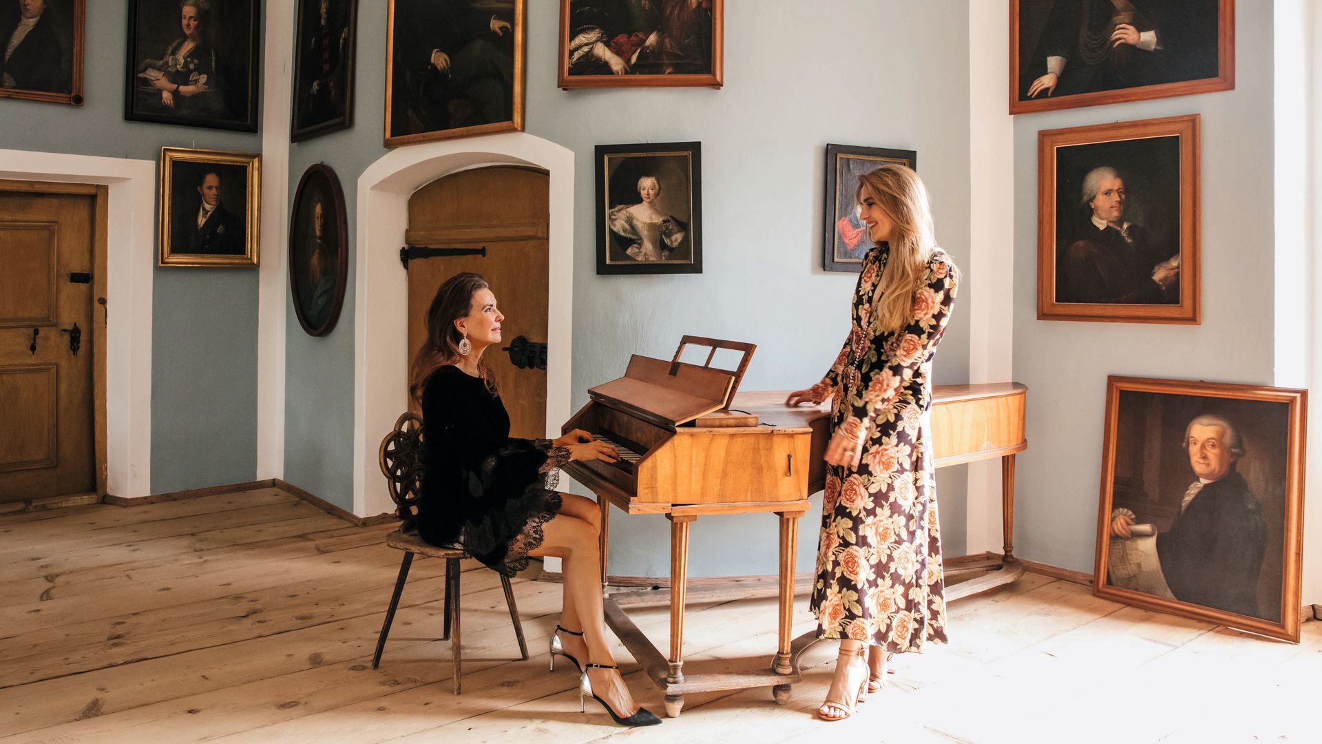 Katrin y Philline en el salón tocando un clave en una sala con retratos de personas históricas en las paredes.
