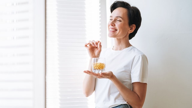 Mujer joven sonriendo con suplemento de colágeno al lado de una ventana