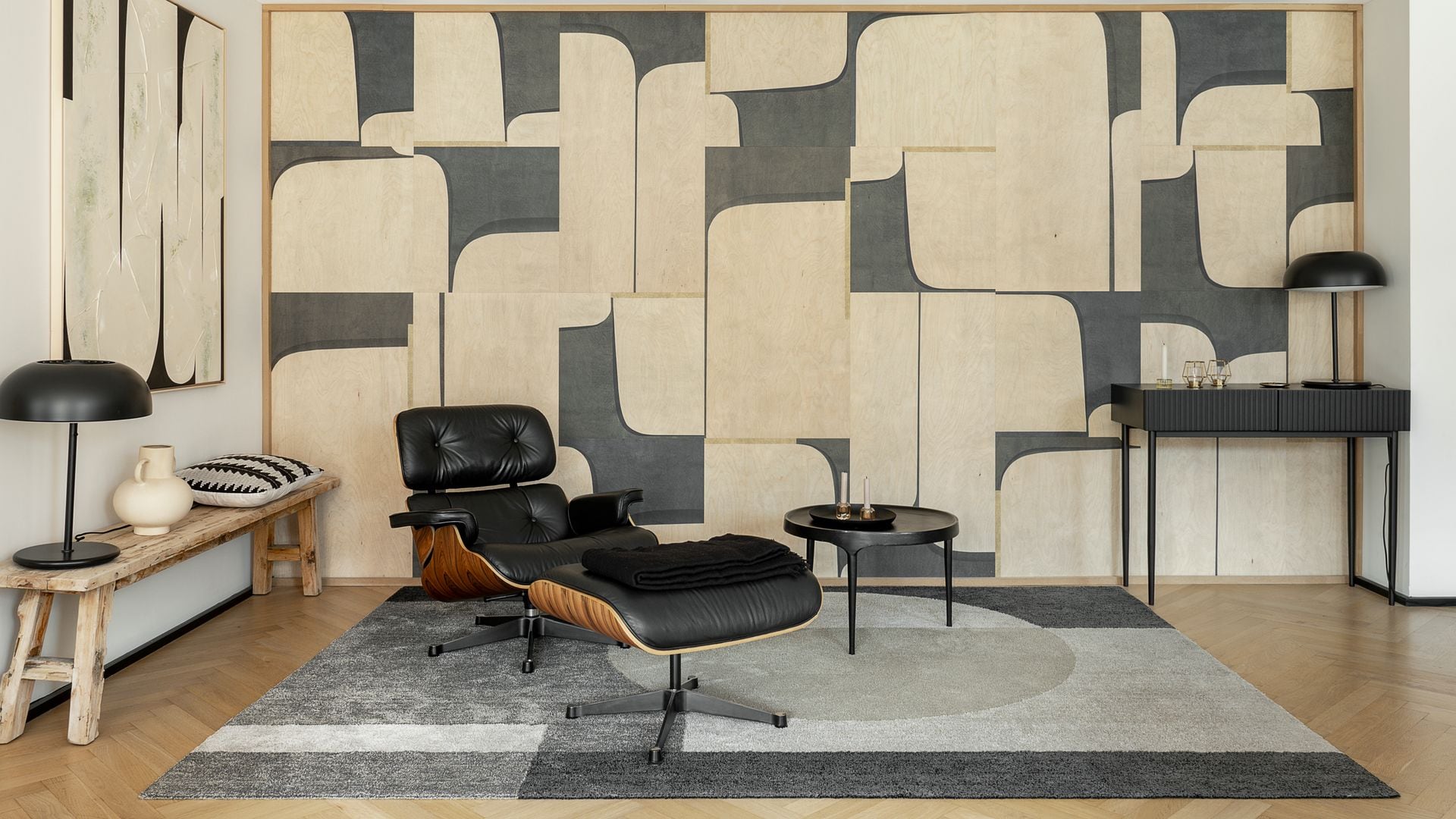 Rincón de lectura con papel pintado, Loungue Chair de los Eames, consola negra, alfombra de motivos geométricos, cuadro en la pared, banco con jarrón, lámpara de sobremesa negra 