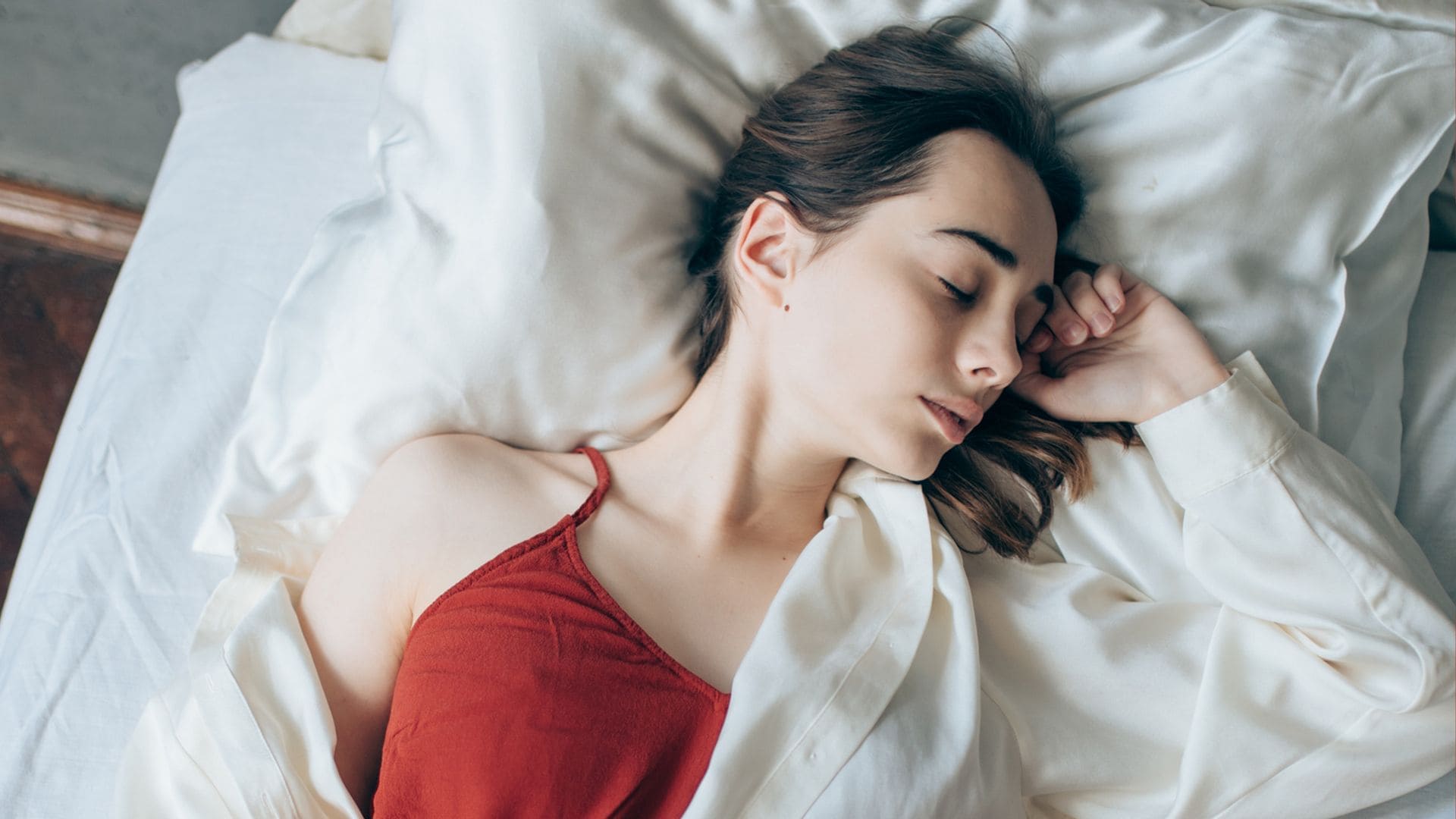 Mujer joven durmiendo sobre una almohada de seda, con camiseta roja y pijama de seda blanco. Está tapada a medio cuerpo con una manta rosa. Plano cenital.