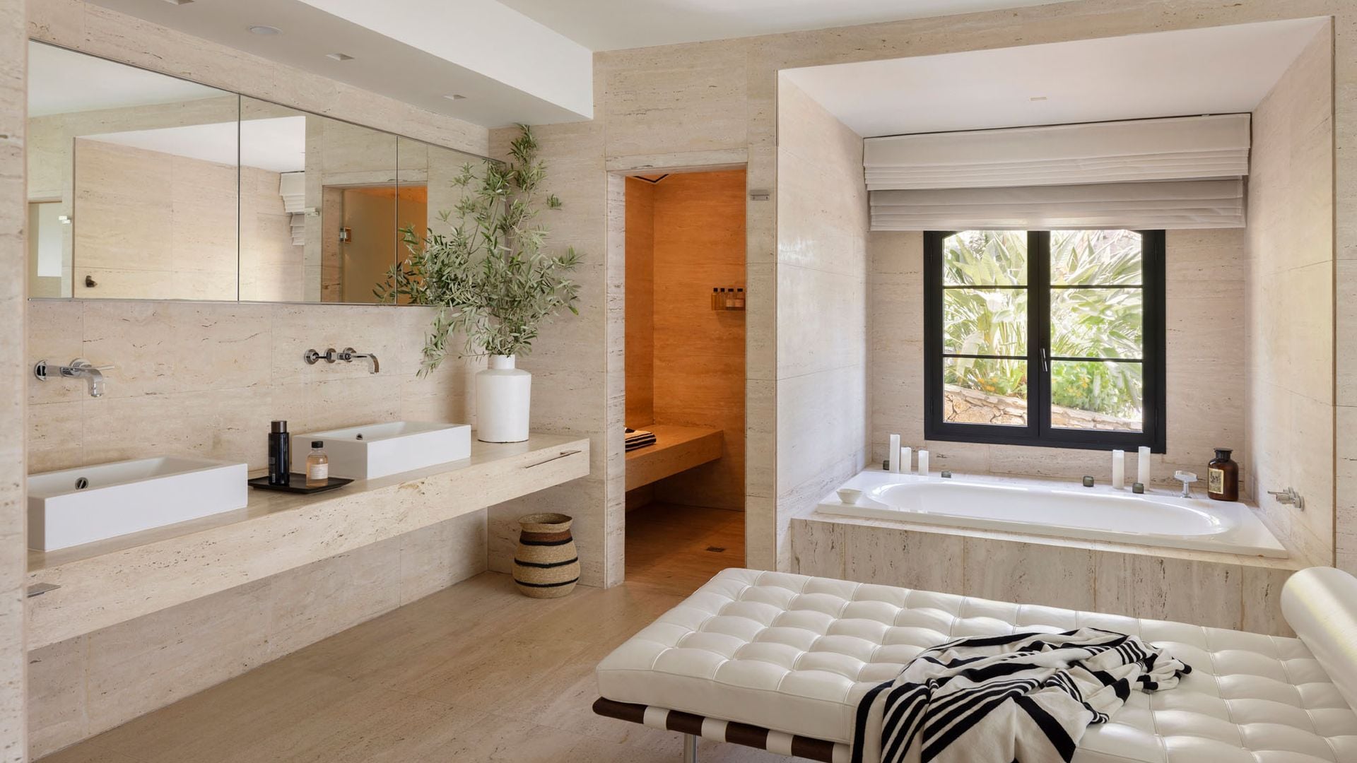 Baño con bañera bajo la ventana, chaise longue de cuero blanco, suelo y encimera de mármol, espejo doble,  doble lavabo