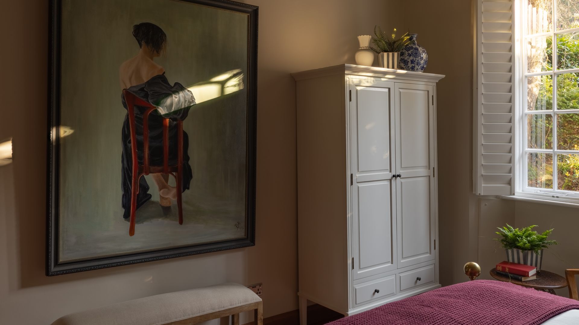 Dormitorio con armario exento, cuadro en la pared de una mujer, banqueta, plaid burdeos a los pies de la cama 
