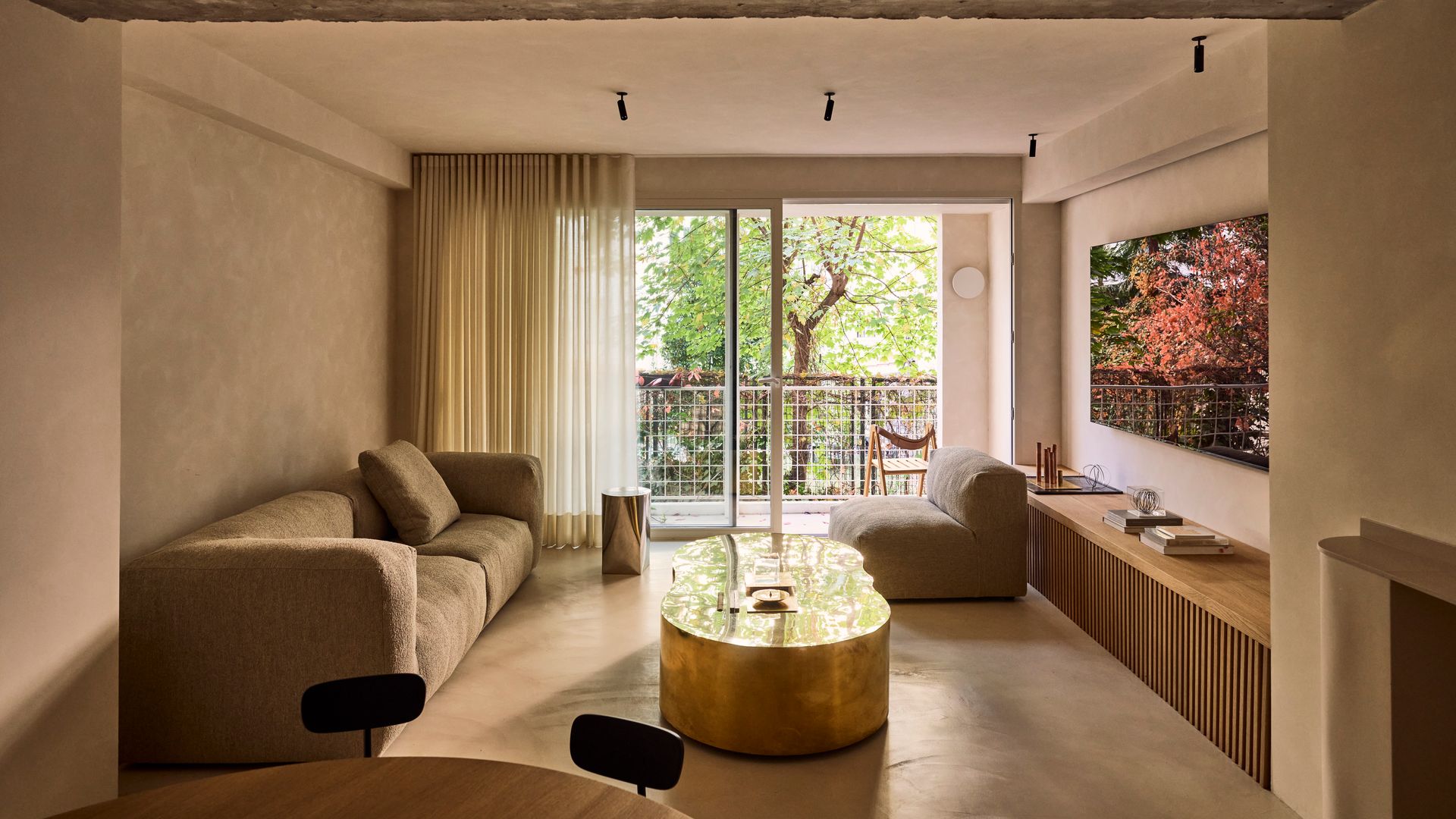 Salón comedor con salida a la terraza con aparador bajo de madera, mesa de centro dorada metalizada, sofá marrón, cortinas blancas, televisión en la pared