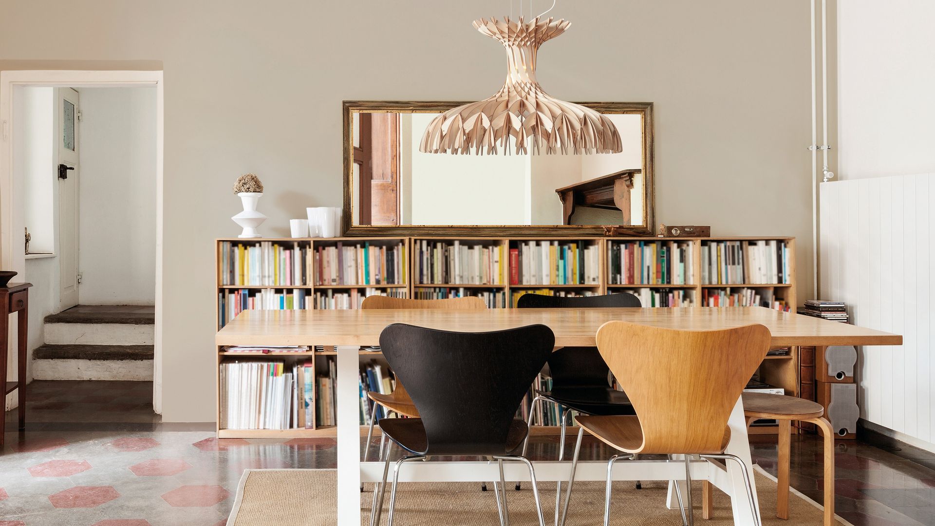 Comedor con lámpara de techo de fibras vegetales, techo de madera blanca, mesa de madera, sillas de madera y negras, espejo, librería a media altura llena de libros