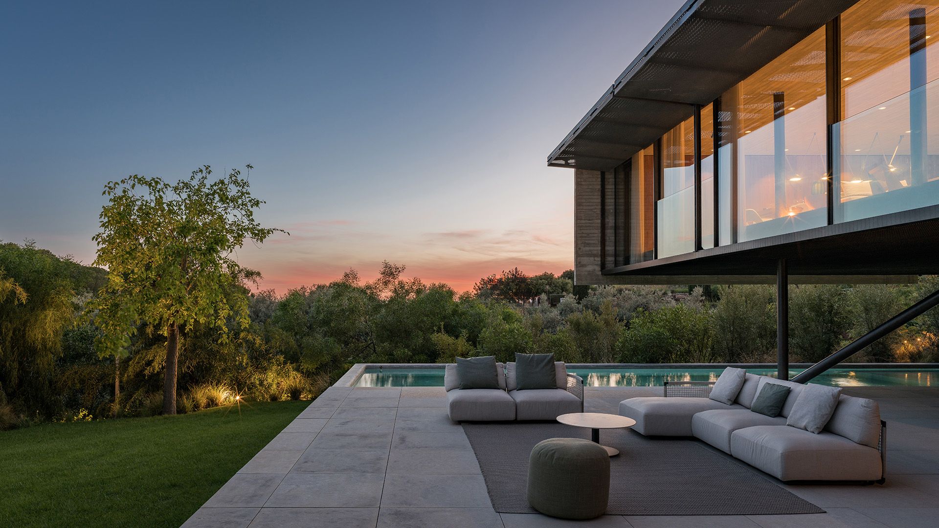 Casa con piscina con fachada de cristal, salón de exterior con sofás y alfombra, césped e iluminación exterior