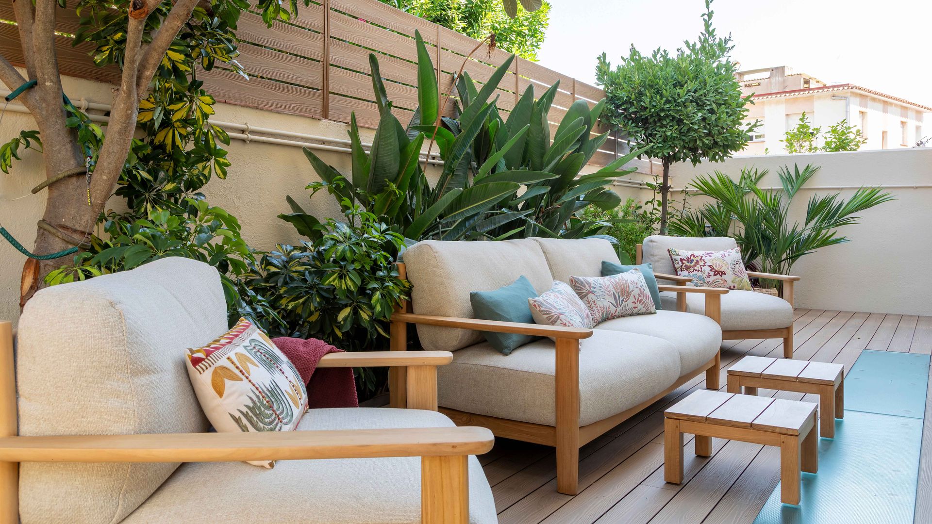 Terraza con suelo de madera, sofás de madera y asientos blancos, mesas auxiliares de madera, muchas plantas en maceta 